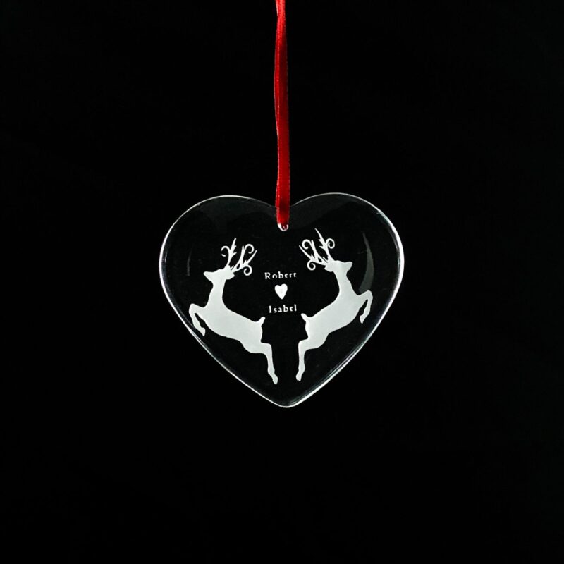 Heart-shaped crystal Christmas ornament with an engraved reindeer design, hanging from a red ribbon.